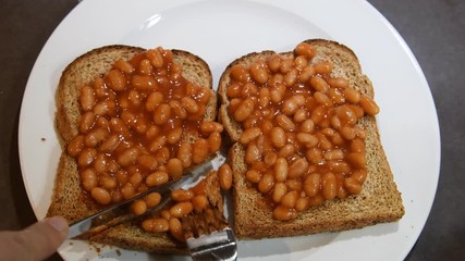 Beans on toast. Hand appearing holding knife and fork and cutting toast with beans on them. Comfort food. Two slices of brown whole meal toast topped with baked beans. Simple British breakfast meal.