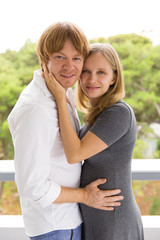 Happy expectant parents hugging and posing outside. Man and pregnant woman standing on balcony, embracing each other and looking at camera. Expecting baby concept