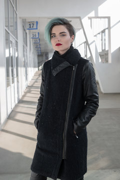 Beautiful girl with short blue fashionable hairstyle in black coat standing in a long white corridor and looking at the camera.