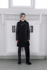 Beautiful girl with short blue fashionable hairstyle in black coat posing on the light background. Head turning right, hands along the body. Full-length photo.