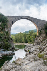 A young hiker contemplates the old medieval bridge of Llierca, in the region of La Garrotxa, Girona, Spain, on a cloudy dramatic day.