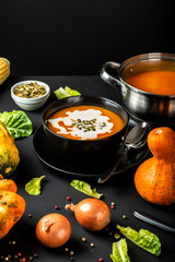 Freshly made pumpkin cream soup with ingredients and saucepan on a table on a black background.