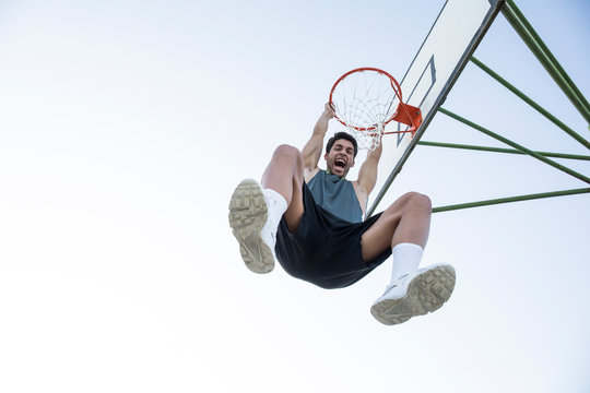 Young man screaming while hanging on basketball hoop and looking at camera