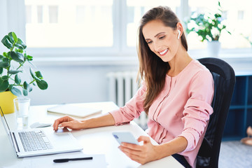 Young businesswoman checking phone while working on laptop in office