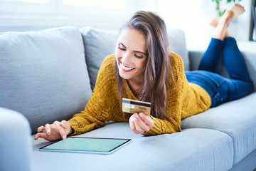 Smiling young woman using credit card and tablet to shop online while lying on sofa
