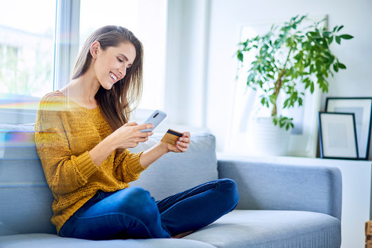Beautiful happy woman paying online with smartphone and credit card while sitting on sofa