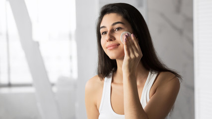 Fototapeta premium Young Woman Removing Makeup Using Cotton Pad In Bathroom