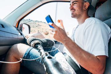 Smiling young man with leg prosthesis sitting in camper van using smartphone