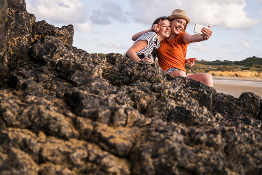 Two girlfriends sitting on rocky beach, taking smartphone selfies