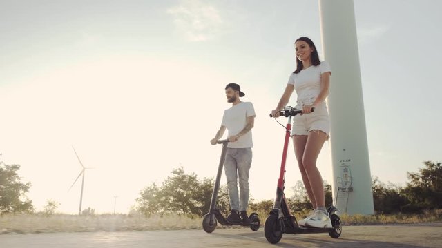 A guy and a young girl at sunset ride on electric scooters on the background of wind power generators. Alternative energy.