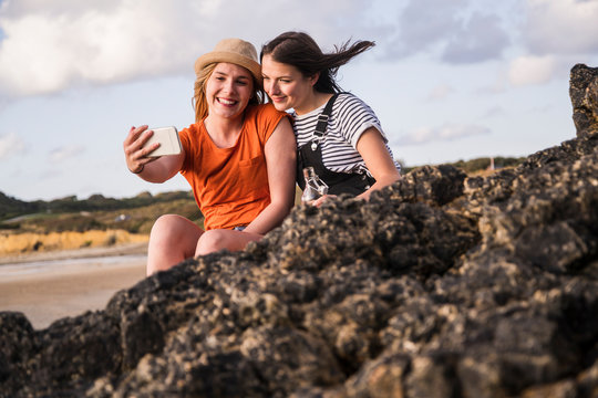 Two girlfriends sitting on rocky beach, taking smartphone selfies