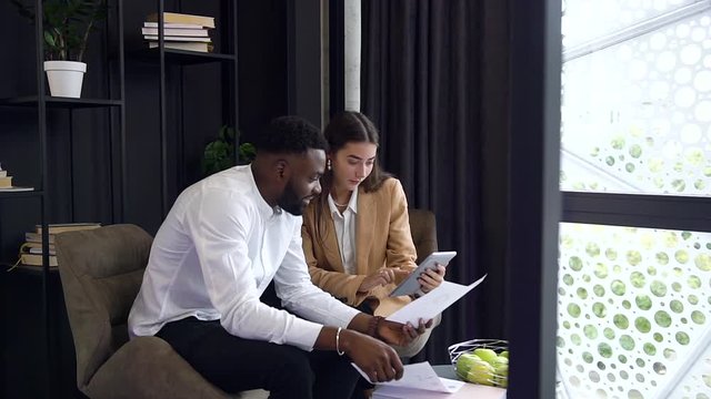 Purposeful pleasant multiracial business people sitting together in the separate office place and using tablet pc and documents ,talking about joint job