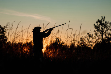 Fototapeta na wymiar Silhouette of a hunter in a cowboy hat with a gun in his hands on a background of a beautiful sunset. The hunting period, the fall season is open, the search for prey.