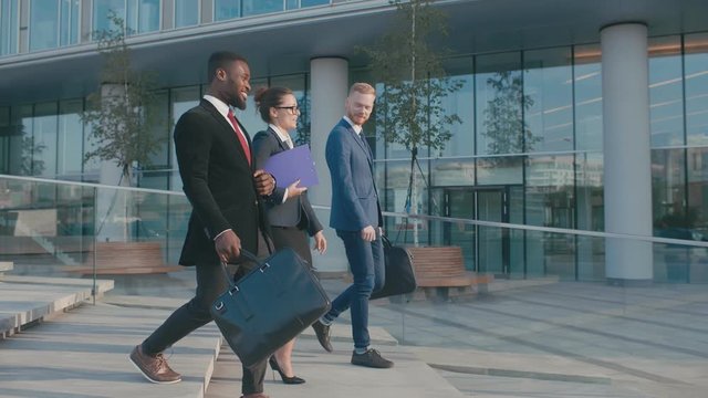 Diverse multi-racial attractive business team with briefcases walk along light modern glass office building facade. Three colleagues partners going down stairs