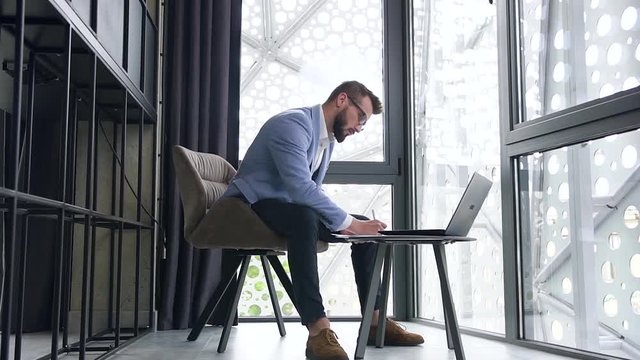Good-looking serious wise bearded businessman sitting in specially designated room and working with documents and laptop