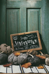 Wool and textile pumpkins for Halloween holidays home decor with autumn leaves, black chalkboard with Happy Halloween lettering on knitted doormat with old wooden door at background.