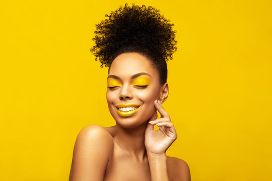 Excitement African American Fashion Model posing against yellow background and touch her face. Head shot portrait . Satisfied Brunette young woman with afro hair style and creative yellow make up
