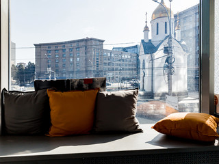 A window with a windowsill on which pillows lie with a view of the city and the attraction of a small chapel with a golden dome in Novosibirsk.
