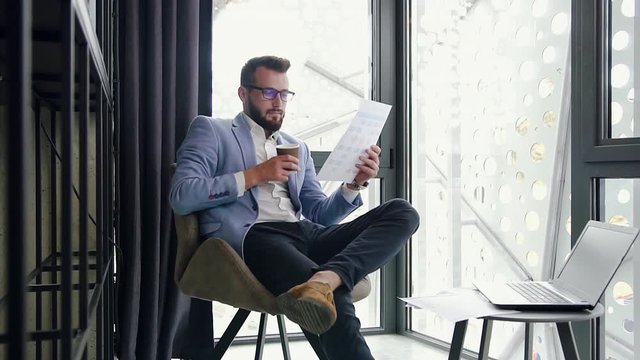 Pleasant confident young bearded businessman reading financial report and drinking coffee from paper cup during his work in the special separate office room