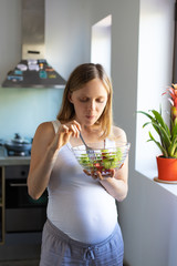 Focused expectant mother enjoying healthy lunch. Pregnant woman standing in kitchen, holding bowl, eating salad. Pregnancy and organic diet concept