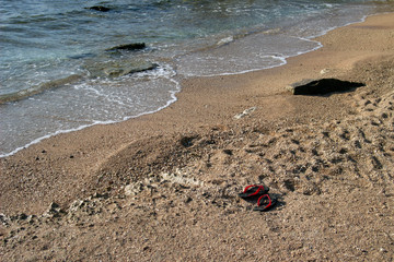 Summer background of hot sand with sea or ocean wave bubbles with copy space for card or advertisement at dawn or sunset. A pair of beach red and black slippers