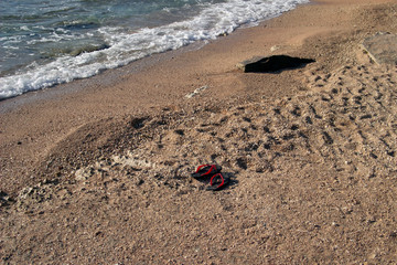 Summer background of hot sand with sea or ocean wave bubbles with copy space for card or advertisement at dawn or sunset. A pair of beach red and black slippers