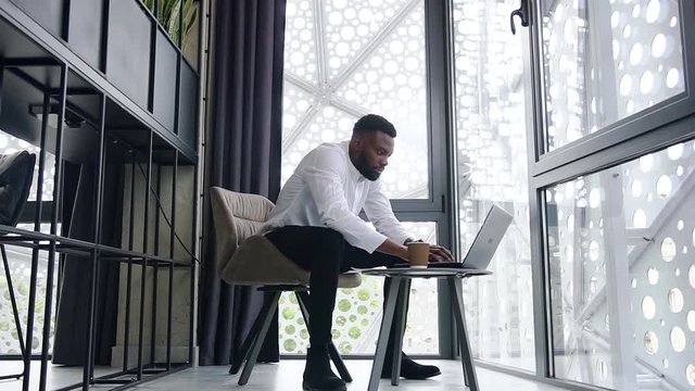 Focused wise bearded african american office worker typing at the computer in separate place in modern office with big window