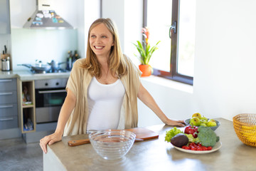 Joyful expectant mother cooking salad in kitchen. Young pregnant woman standing at table with bowls of vegetables, smiling, looking away. Pregnancy and nutrition concept