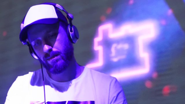 DJ performs on nightclub stage. Circular fly around bearded disc jockey playing live show at disco in slowmotion. Night dance club electronic music party. Man mixing music on sound console.