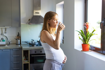 Pensive expectant mother drinking water and looking through window. Pregnant woman with glass of...