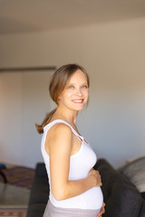 Happy expectant mother posing in living room. Cheerful pregnant young woman holding baby bump, smiling, looking away. Maternity concept