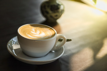 Close up of white cup of hot coffee latte with milk foam art with plate and spoon on wooden table with morning sunlight and shadow.