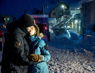 Happy young snowboarder couple, walking in the evening and cuddling at winter resort in snowfall