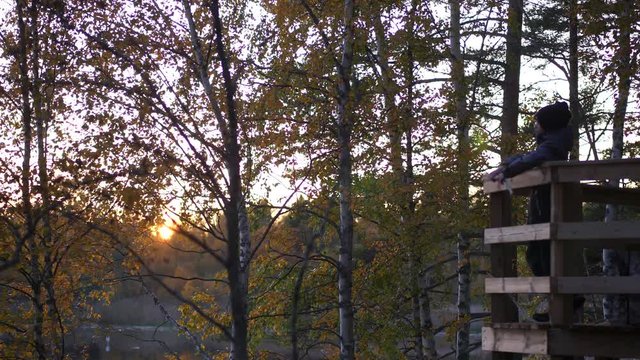 Young boy in a lookout tower enjoying beautiful view of lake and yellow trees, cold autumn sunset