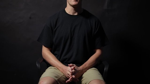 Shot of a man sitting in a chair with his fingers unlocked, rubbing his hands together. Shot in a studio with a black background.