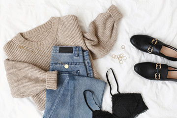 Blue jeans, beige knitted sweater, black lace bra, black loafers or flat shoes lying on bed on...