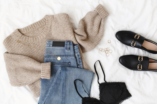 Blue jeans, beige knitted sweater, black lace bra, black loafers or flat shoes lying on bed on white sheet. Overhead view of women's casual day outfit. Trendy women clothes. Flat lay, top view.