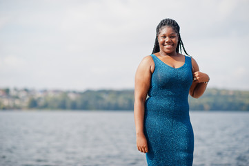 African american dark skinned plus size model posed in a blue shiny dress against sea side.