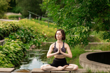 Young woman doing yoga. Girl sitting in the lotus position in the park near a small decorative lake.