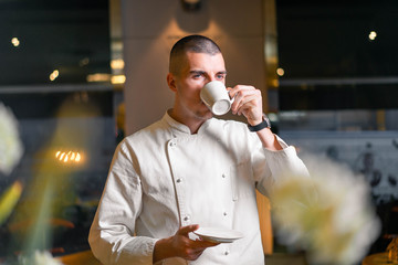 Young handsome cook chief drinking morning coffee in modern luxury restaurant interior.