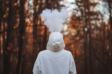A young man in a white hoodie exhales a cloud of steam smoke up against the background of an autumn...