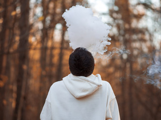 A young man in a white hoodie exhales a cloud of steam smoke up against the background of an autumn Park