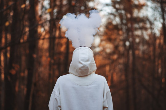 A young man in a white hoodie exhales a cloud of steam smoke up against the background of an autumn Park
