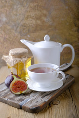 A white cup and a white teapot with tea, a jar of honey and fresh ripe figs on the cutting board on the table.