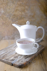 A white cup of hot tea and a white kettle on a wooden table.