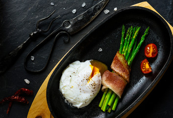 Poached egg with asparagus wrapped in fried bacon, with cherry moid in a pan on a wooden stand on a black stone background with a meat fork and knife