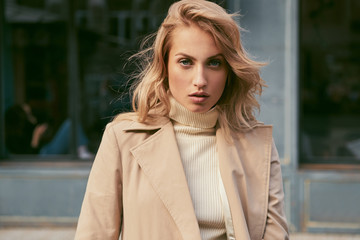 Beautiful serious blond girl in trench coat confidently looking in camera on city street