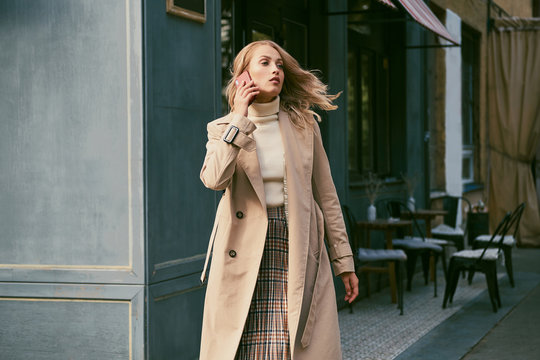 Attractive casual blond girl in stylish trench coat talking on cellphone on street