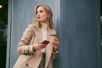 Attractive casual blond girl in stylish trench coat with coffee to go and cellphone thoughtfully...