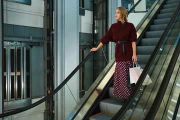 Beautiful stylish blond girl with shopping bags thoughtfully looking away on escalator in shopping mall